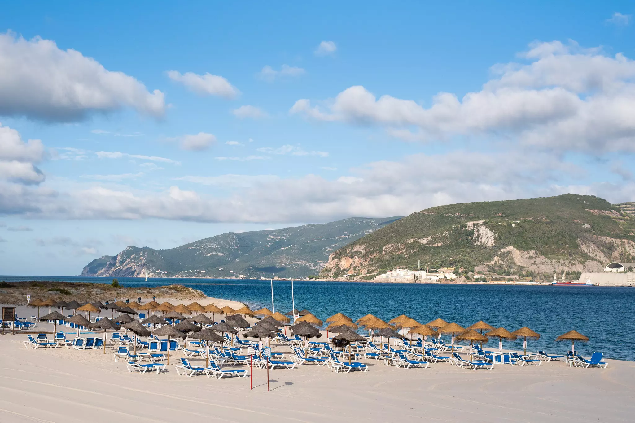 Many grass umbrellas and blue loungers on the sand with blue ocean and hills in the distance on a sunny day.