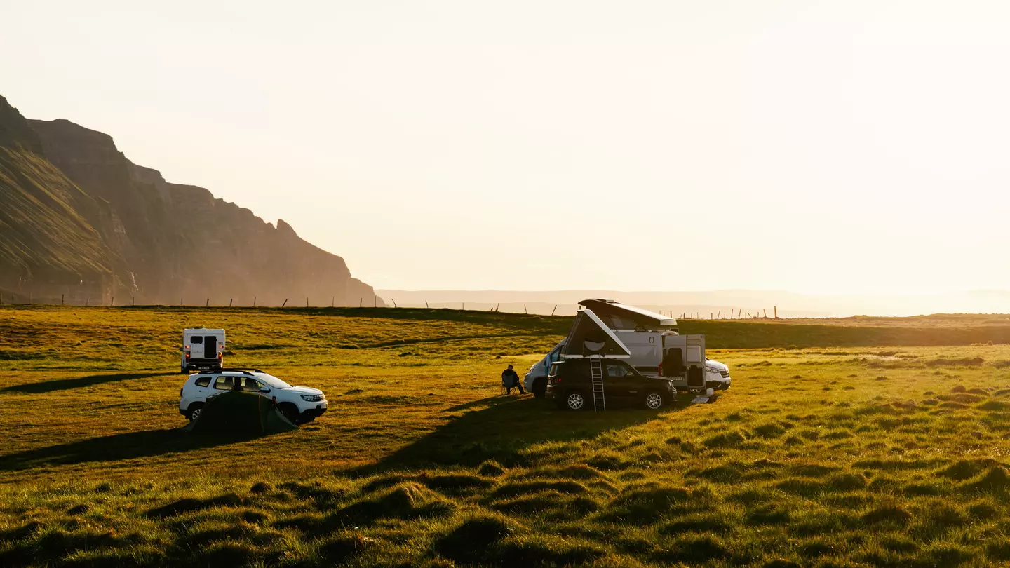 Three car camps on green turf with rocks and a beach in the background at sunset