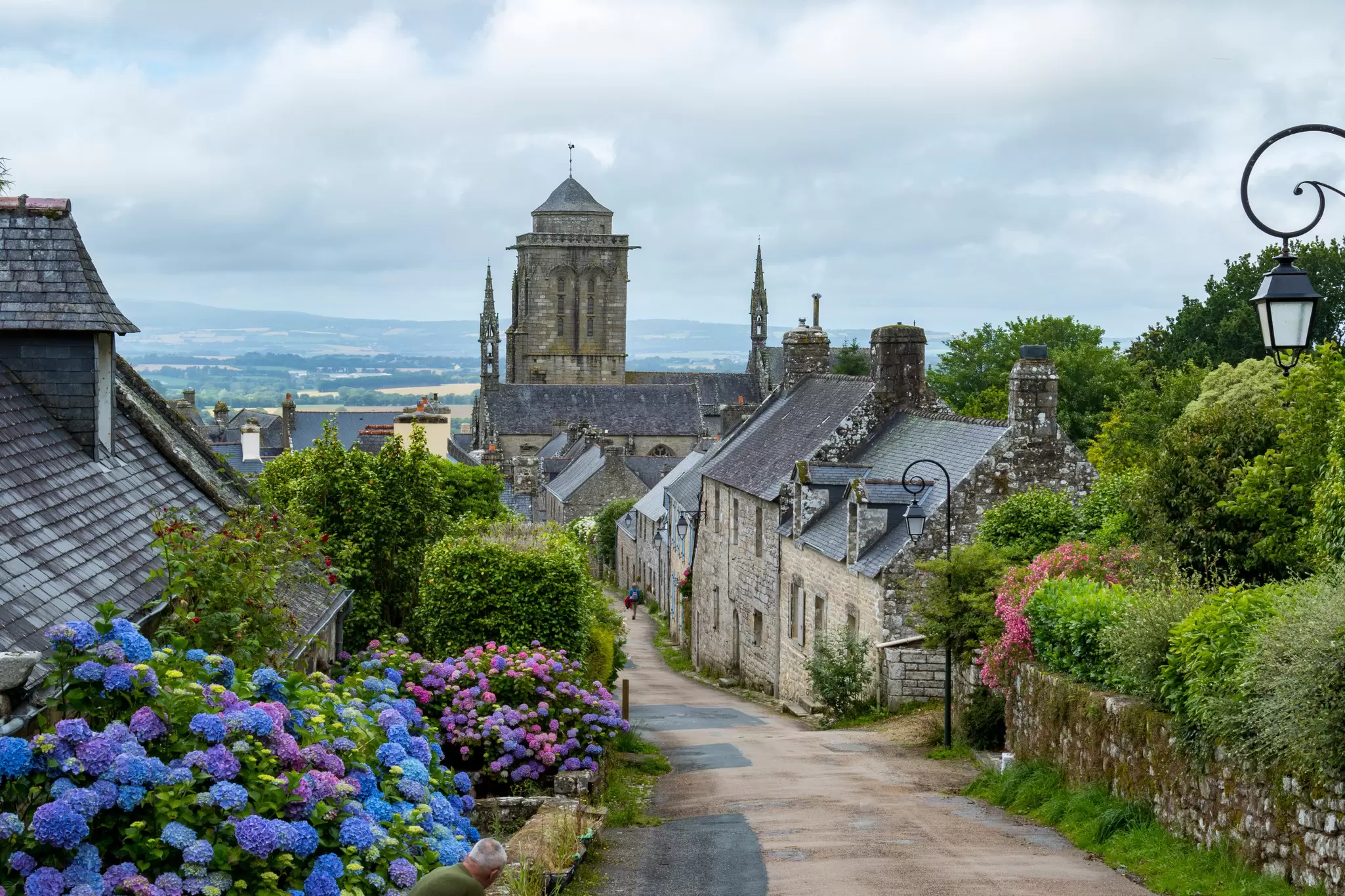 A road through a stone village, with purple flowers blooming on a bush in the foreground.