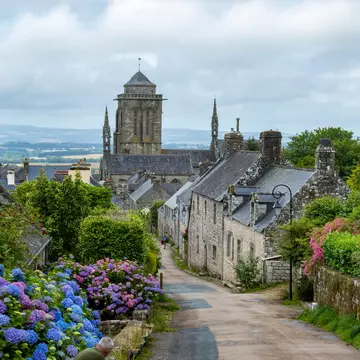 A road through a stone village, with purple flowers blooming on a bush in the foreground.