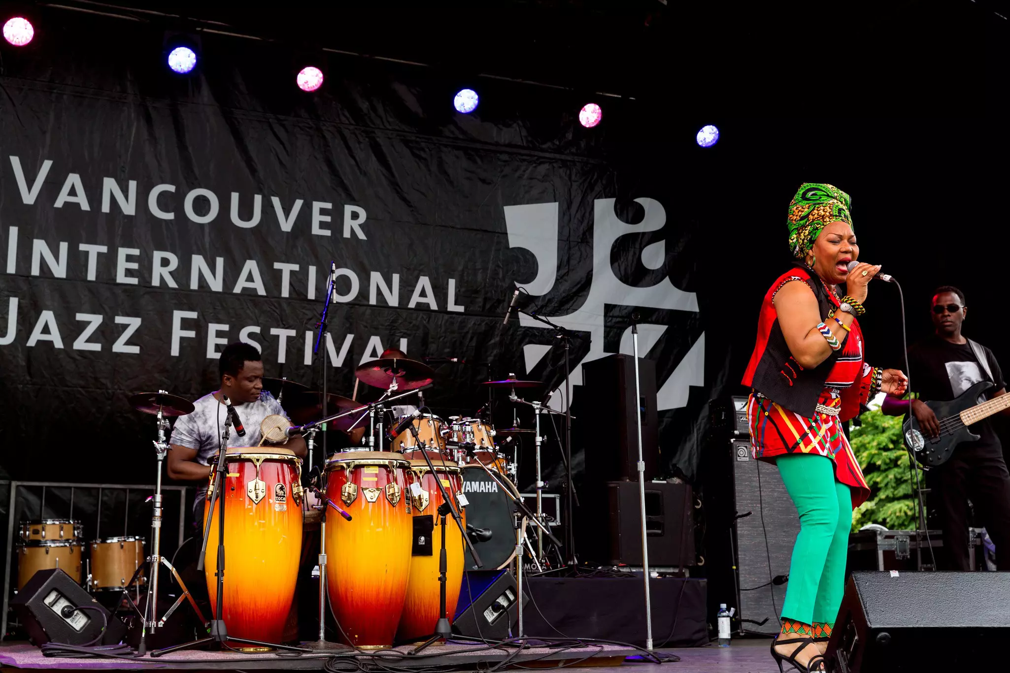 A woman wearing brightly colored clothing and a patterned head wrap performs on stage with her band at Vancouver Jazz Festival.