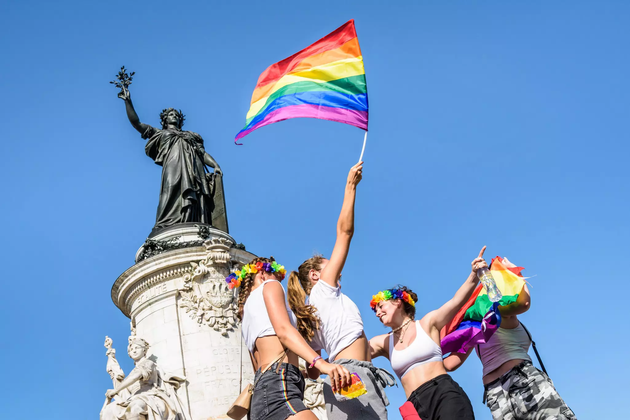 Three young women, one waving a rainbow flag high, dance in the sun at the foot of the statue of Marianne on the place de la Republique, where the Gay Pride parade ended 