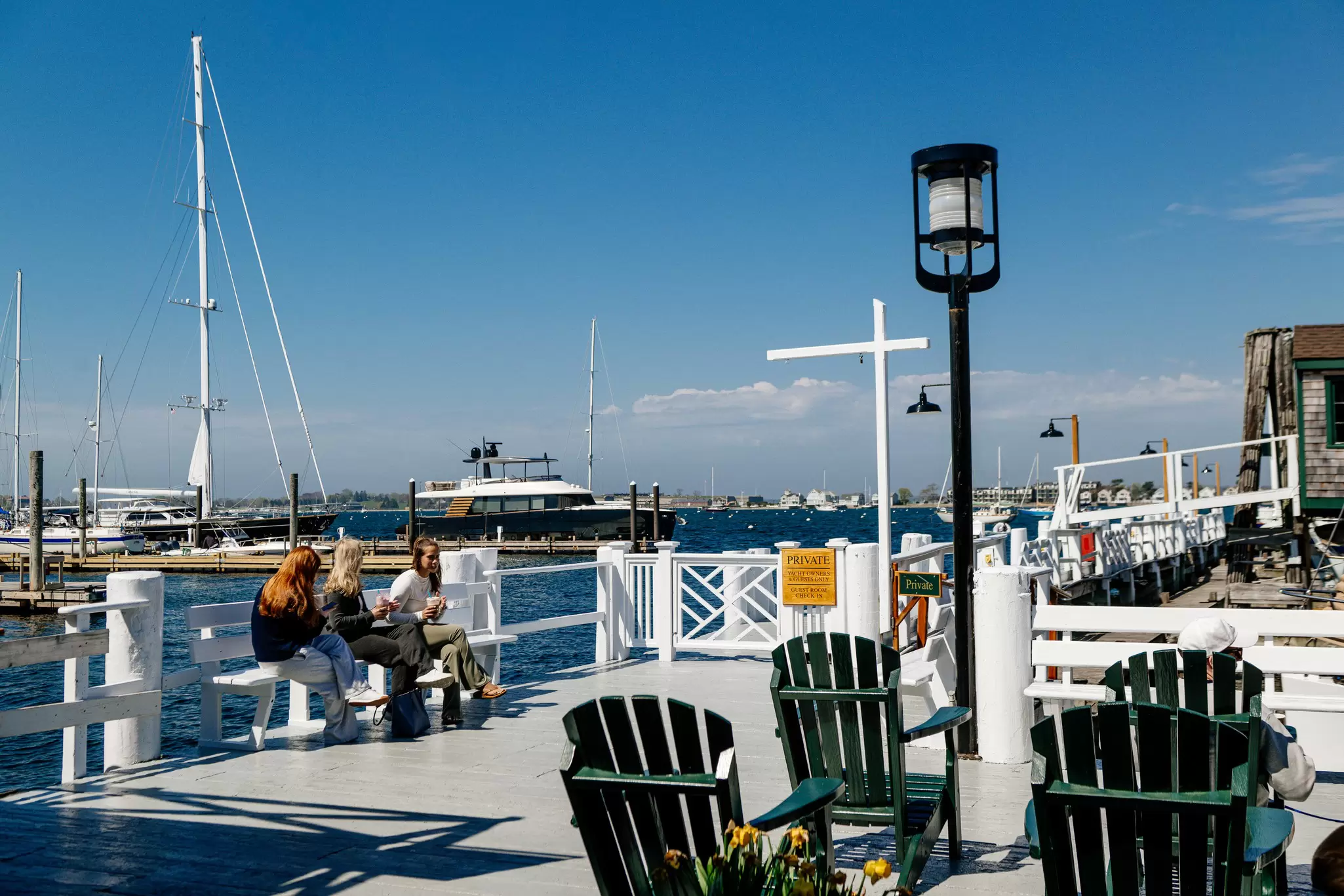 Three people sitting at Bowen's Wharf, Newport, Rhode Island with green deck chairs to the right and boats moored behind