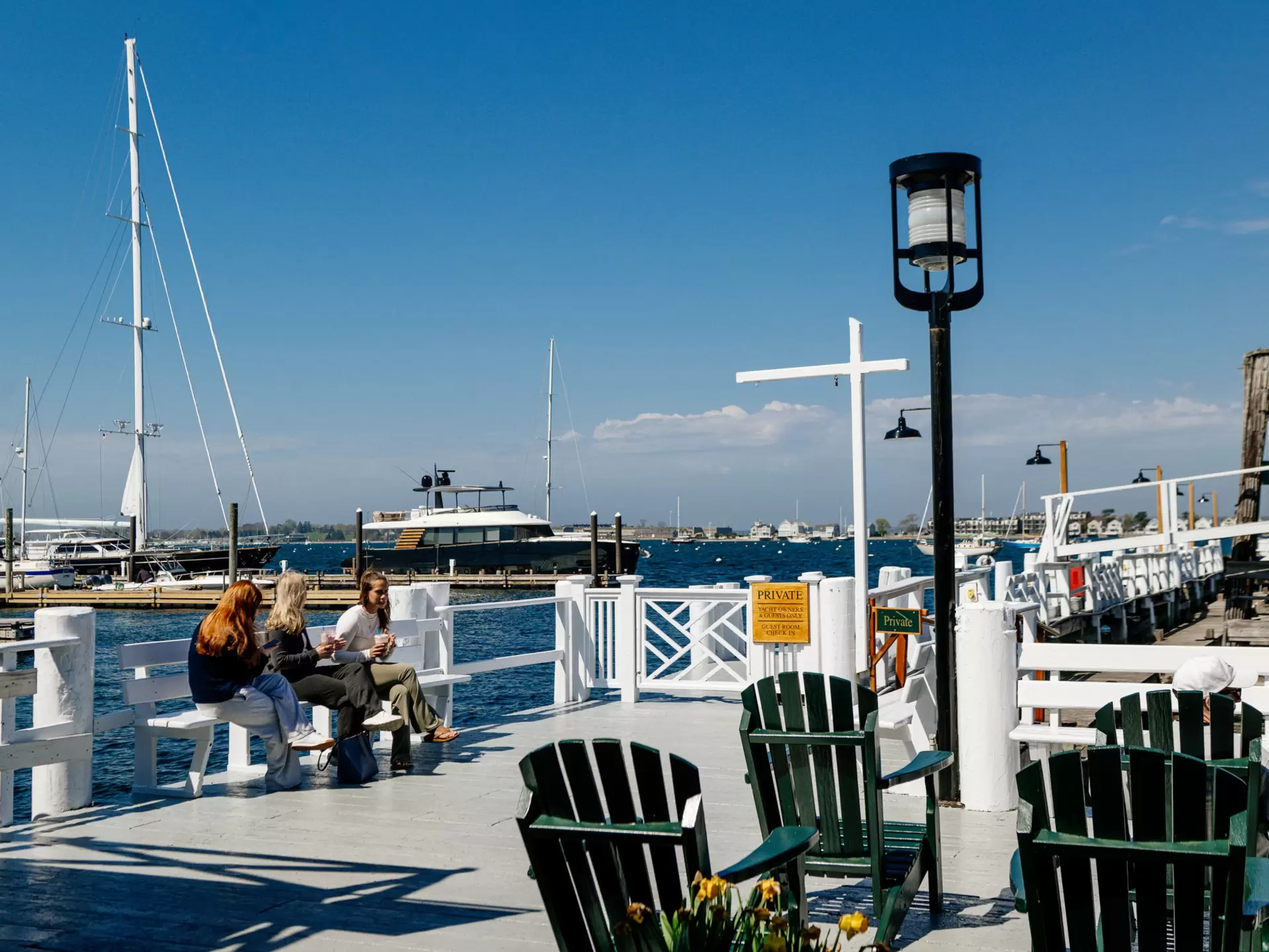 Three people sitting at Bowen's Wharf, Newport, Rhode Island with green deck chairs to the right and boats moored behind