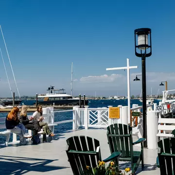 Three people sitting at Bowen's Wharf, Newport, Rhode Island with green deck chairs to the right and boats moored behind