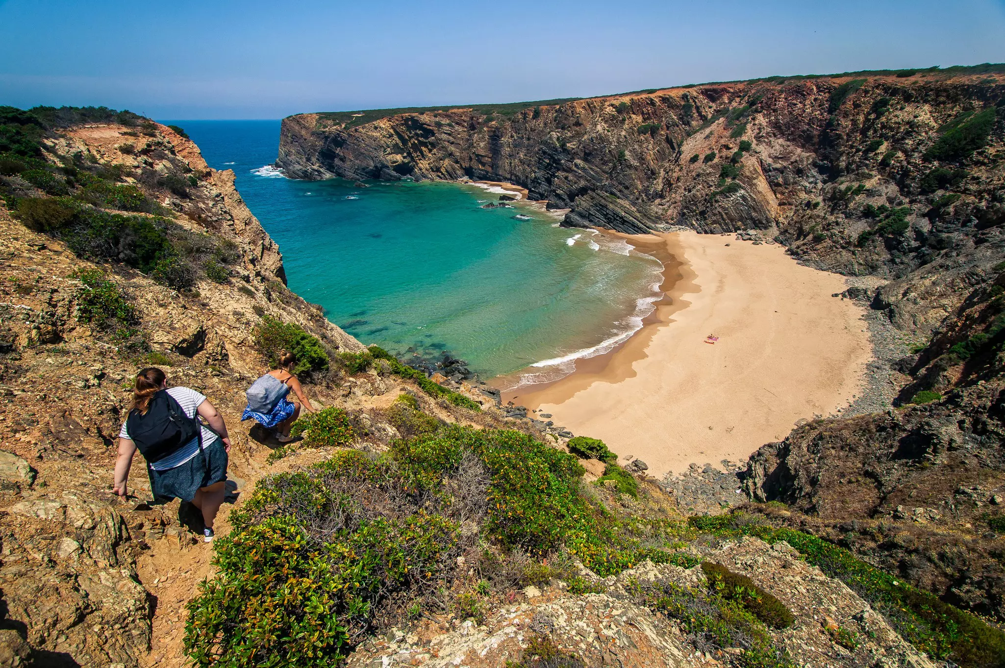 Two hikers scramble down a cliff path leading to a sandy beach