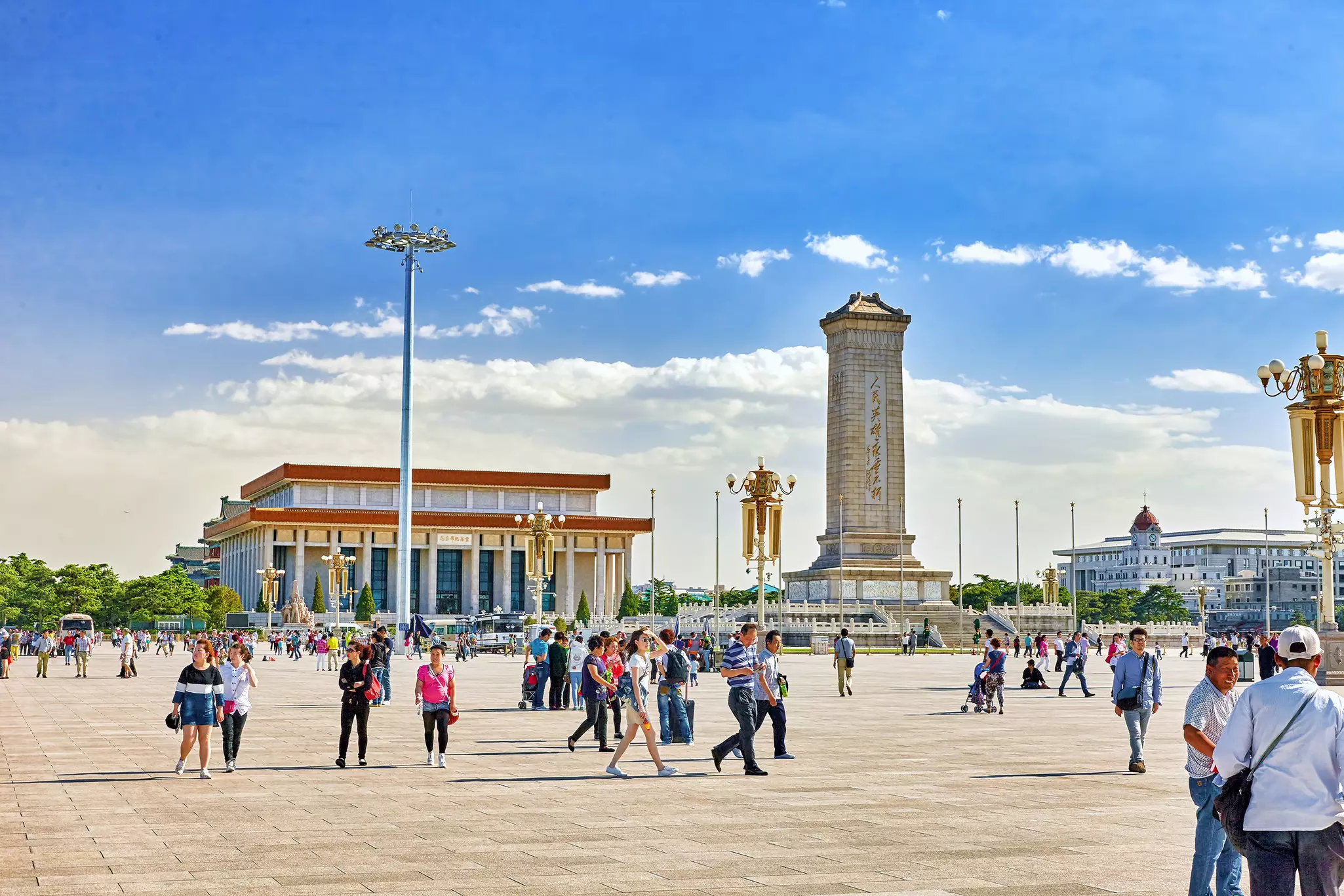 People walk through a large city square lined with large buildings and a tall stone tower.