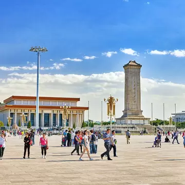 People walk through a large city square lined with large buildings and a tall stone tower: Monument to the People's Heroes on Tiananmen Sq, Beijing.