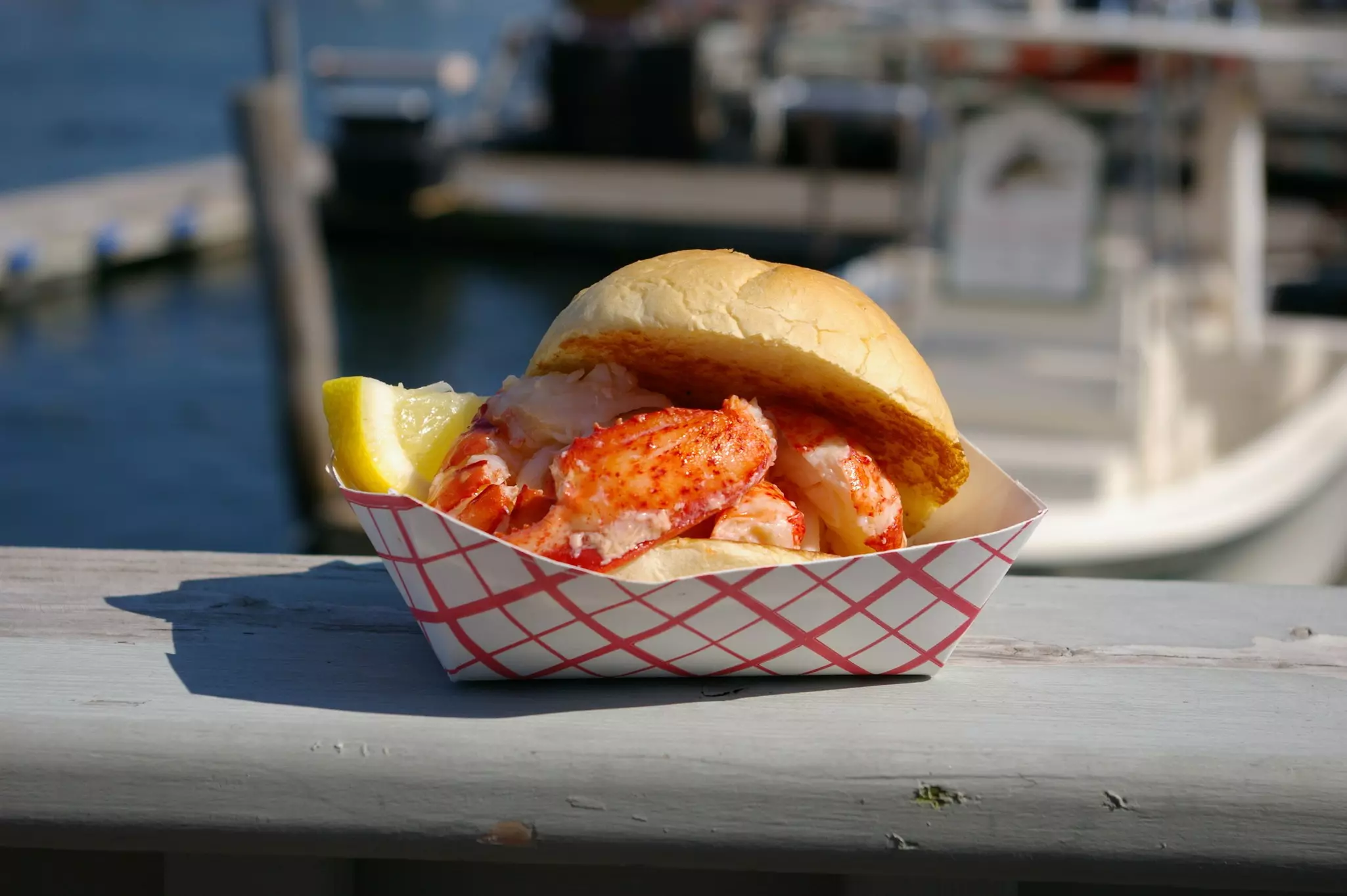 Lobster on a hamburger bun with a lemon wedge in a red-and-white checked cardboard dish sitting on the banister of a dock, with blurry boats in the background