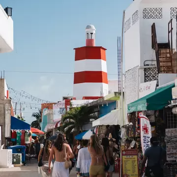 People in swimwear walking down a shop-lined pedestrian street toward a red-and-white striped tower under blue sky.