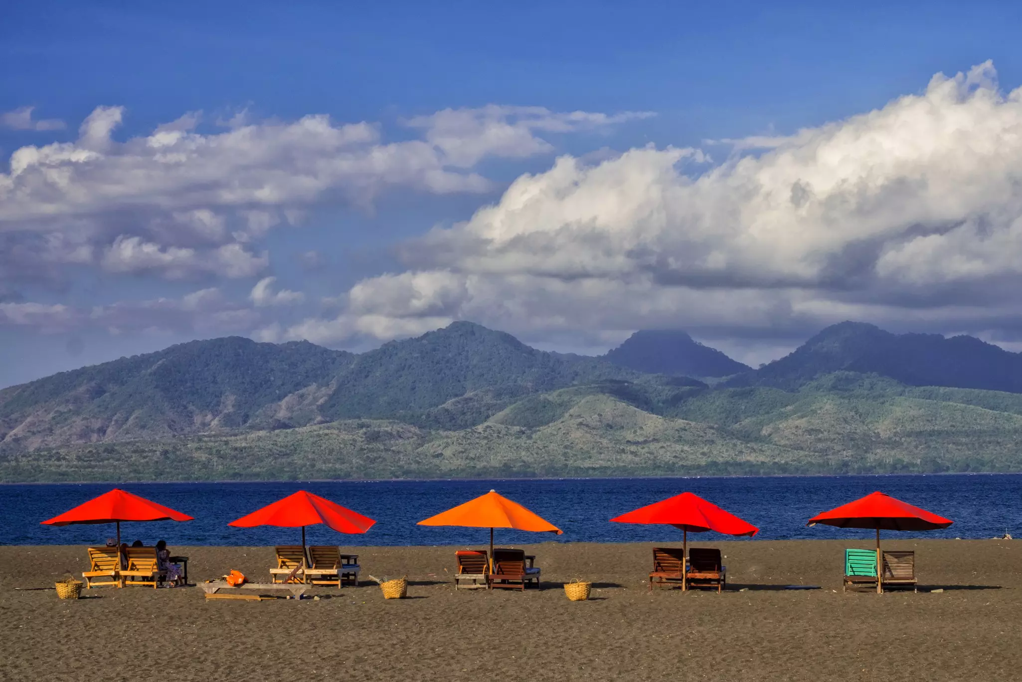 Panoramic view of beach with red and orange umbrellas above wooden lounges with water and forest-covered mountains in the distance.