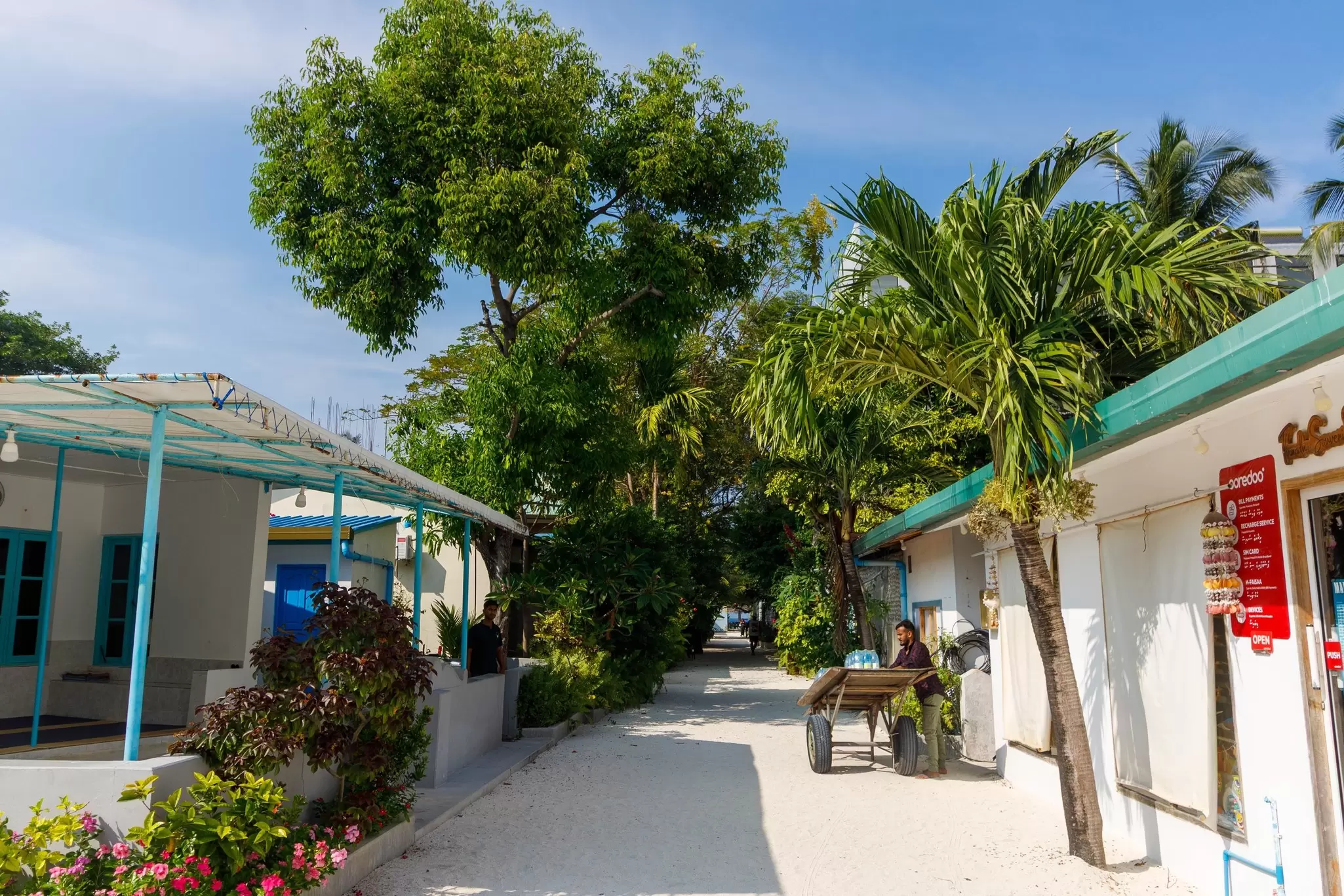 A narrow pathway between homes and trees on the island of Fulidhoo, Maldives. A man sells water bottles from a cart. 