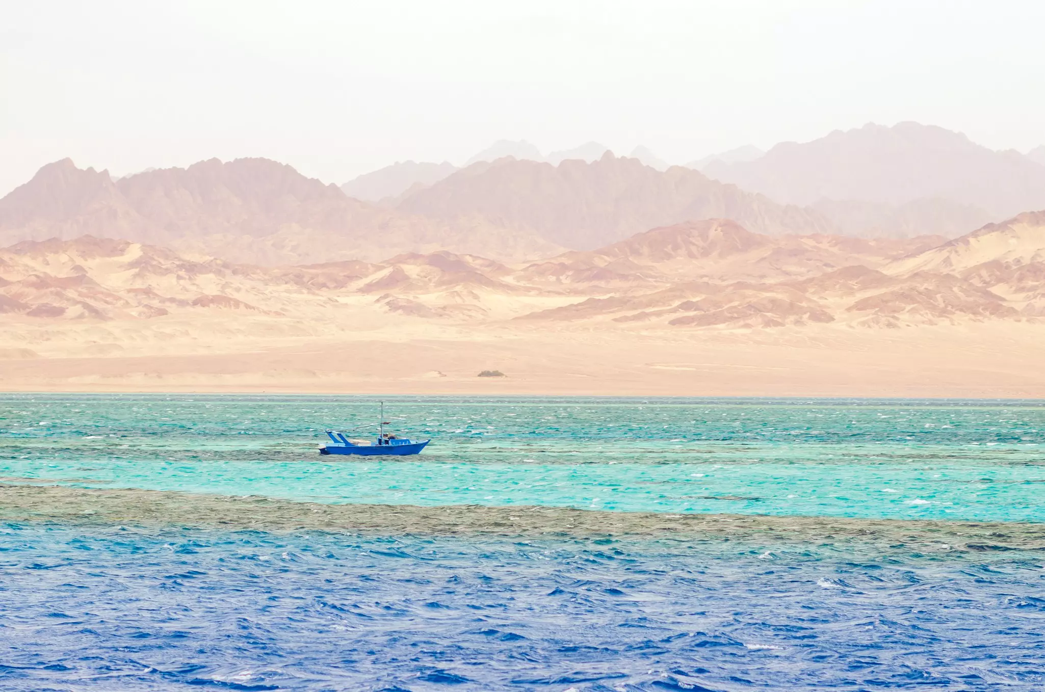 A small boat sails through an ocean striped in turquoise and deep blue. Sandy desert mountains rise in the distance.