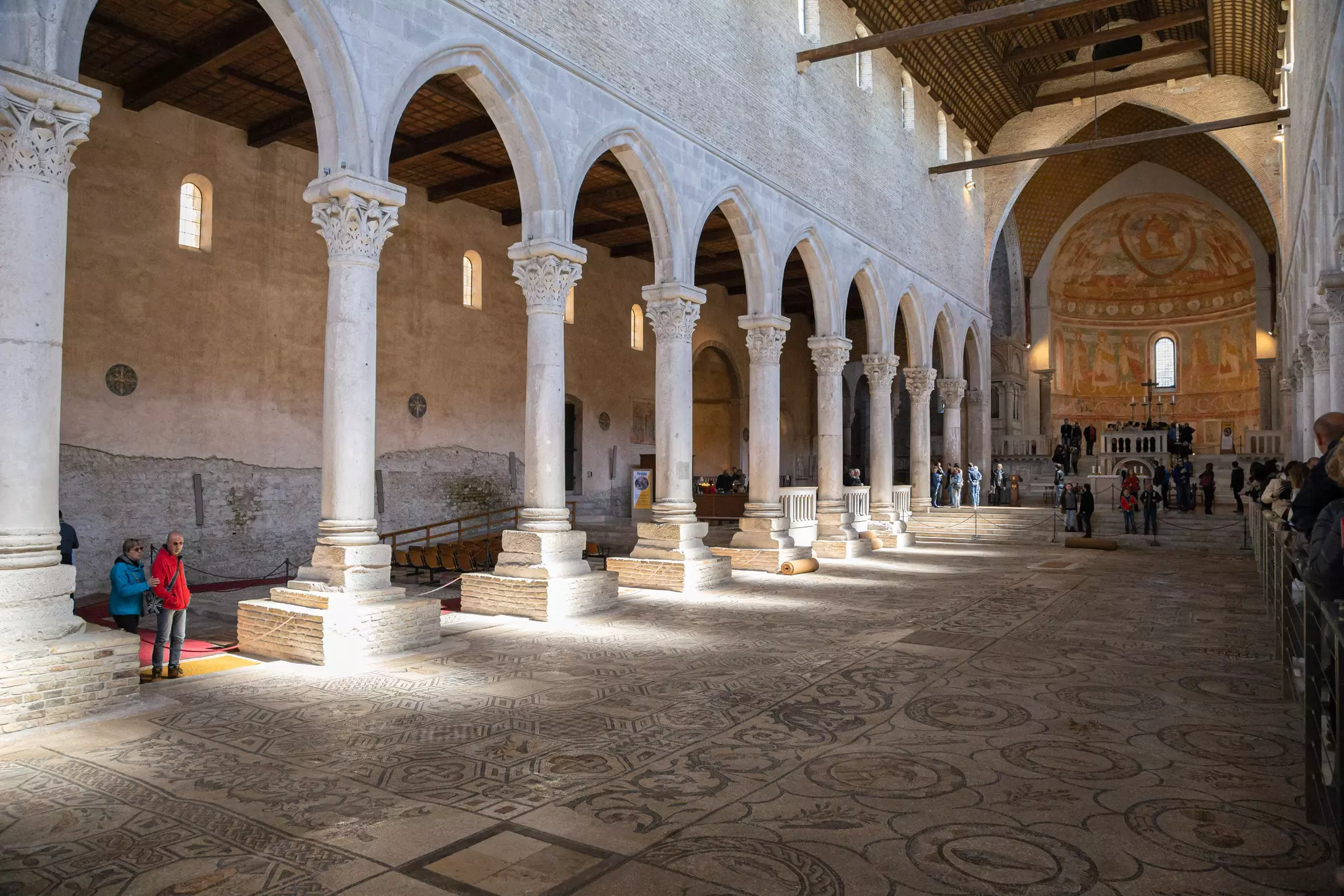 People stand in the nave of a church, supported by columns that frame Gothic arches. A huge mosaic forms the floor of the church, hit sunlight from windows above.