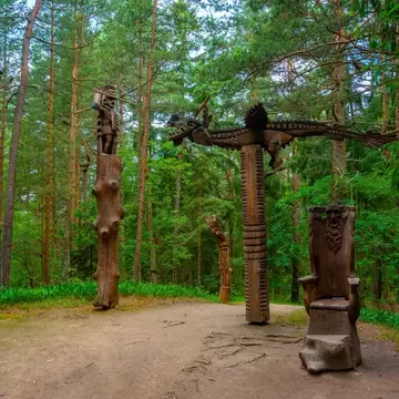 Tall carved wooden figures and a wooden throne on a sandy forest clearing among pine trees.