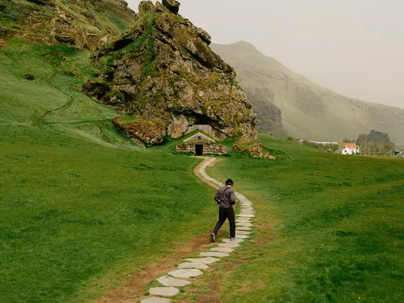 Man walking a stone path among green grass toward a hillside with a manmade cave entrance on an overcast day.