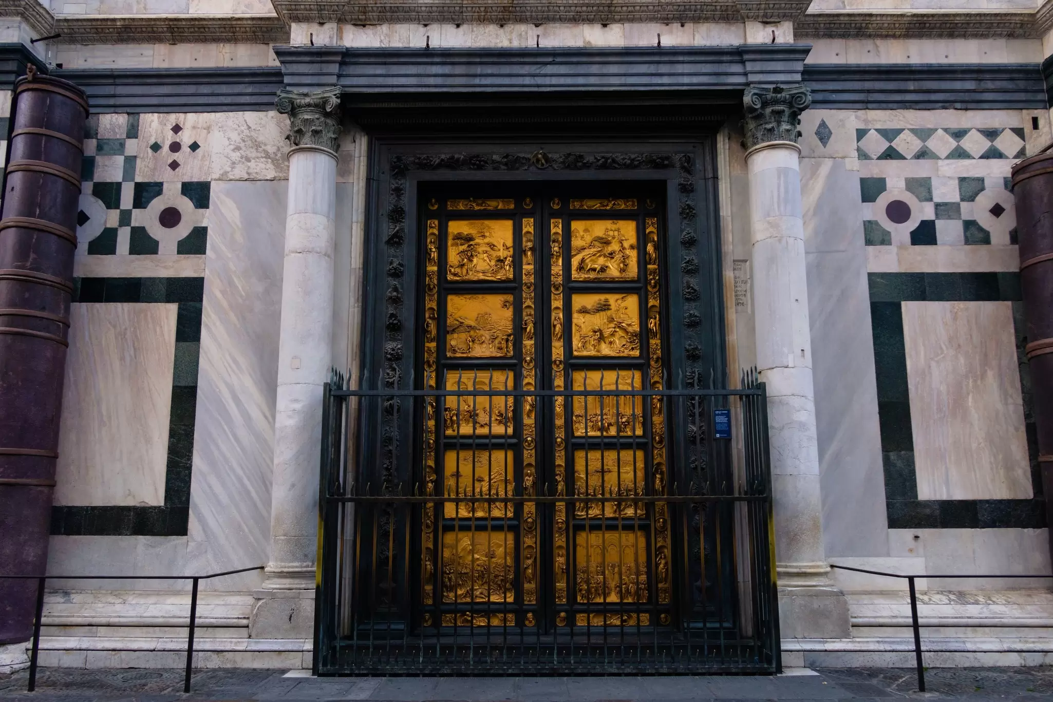 A set of golden doors on a stone building, each with 5 plaques that tell a visual story, behind a gate