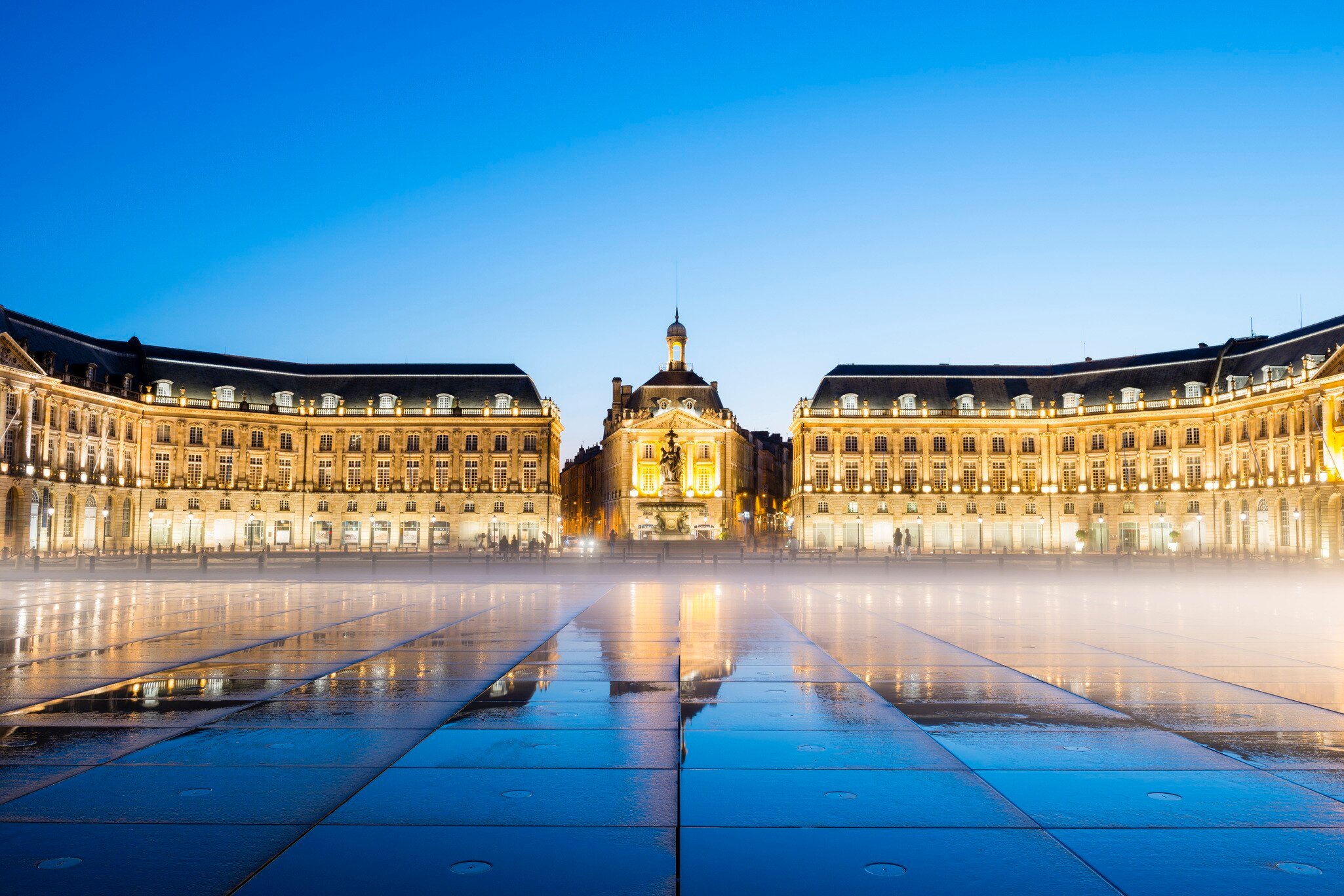 A large city square at dusk. Grand palace-like buildings line one side as a mist hangs just above the ground, which is wet and reflecting light.