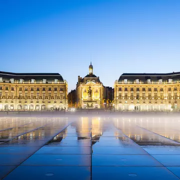 A large city square at dusk. Grand palace-like buildings line one side as a mist hangs just above the ground, which is wet and reflecting light.