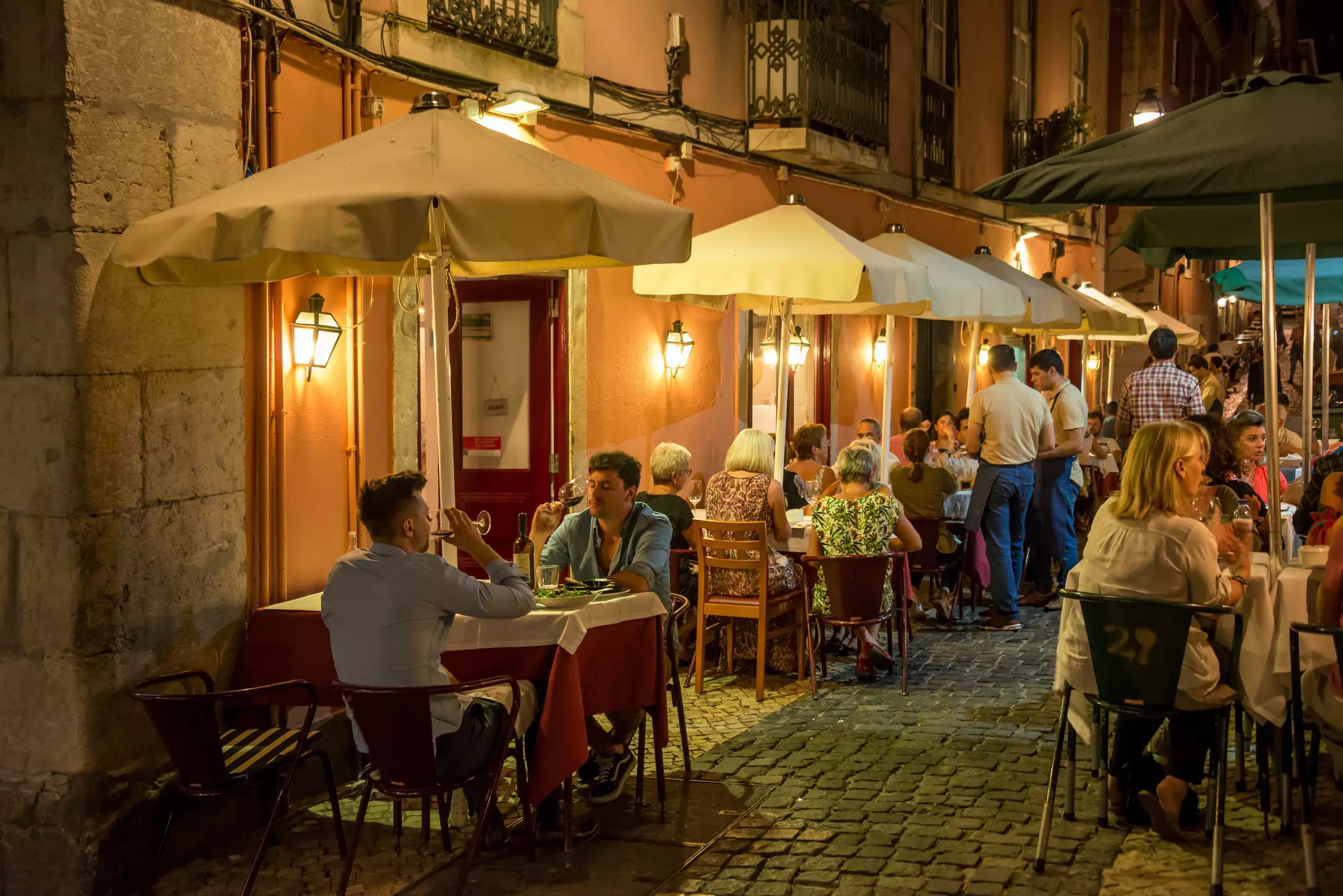 People sit at tables at an outdoor cafe along a cobblestone alley at night.