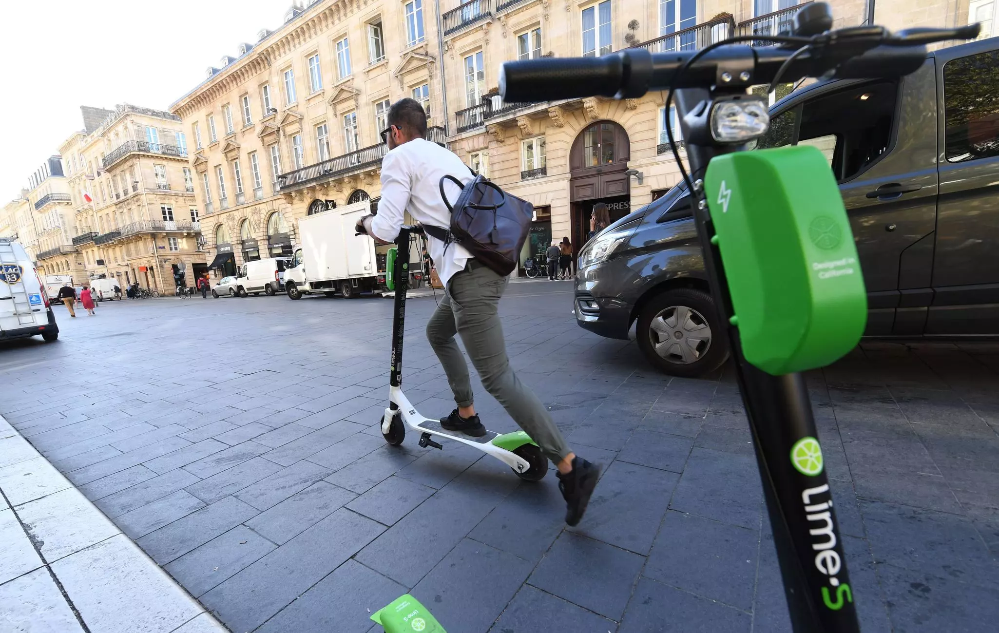 A man pushes off on a dockless e-scooter on a street in Bordeaux, France. Another parked scooter is visible in the foreground.