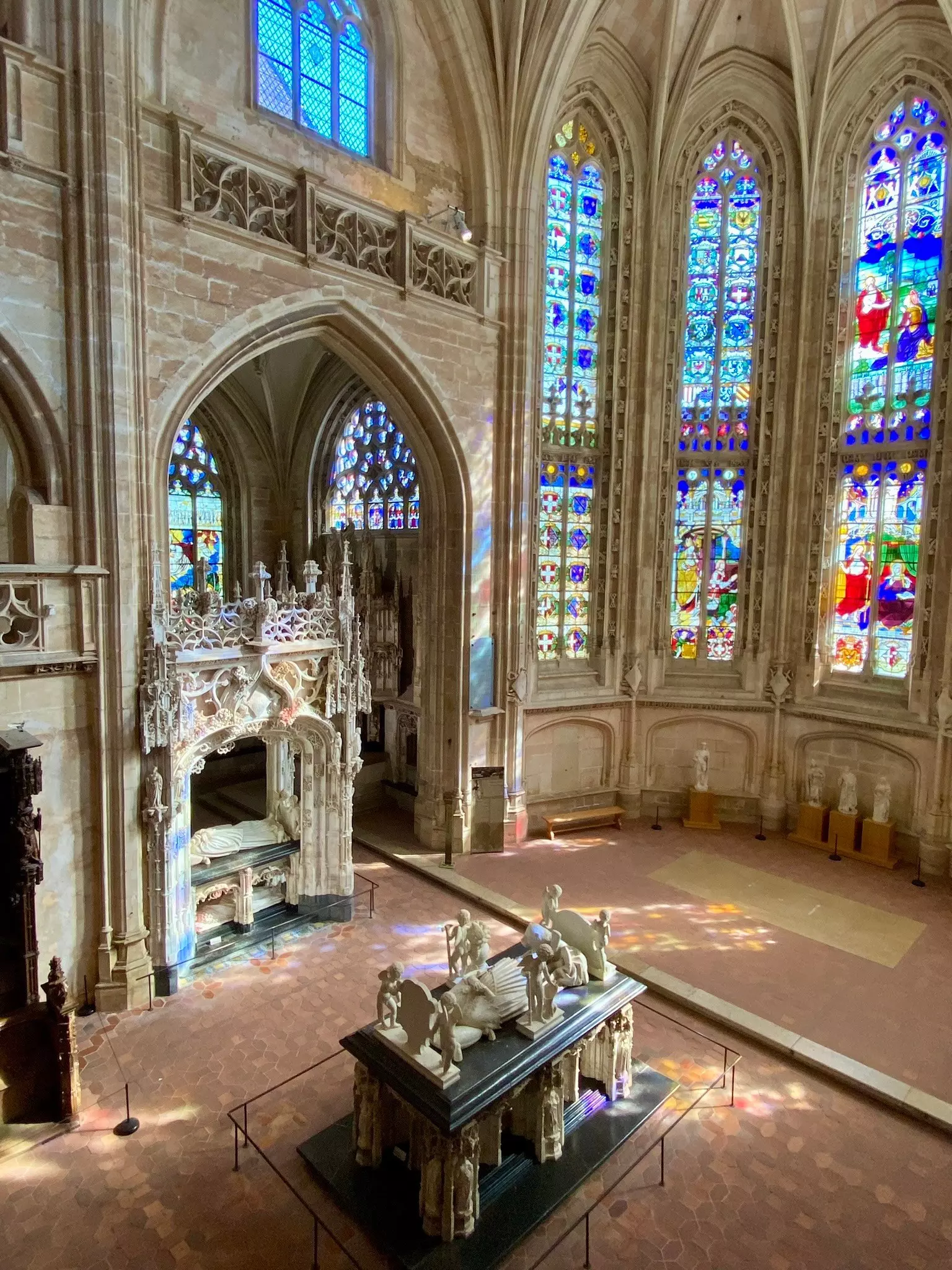 Light floods through a stained-glass window onto marble statues in a large Flaboyant Gothic monastery in Bourg-en-Bresse, France. 