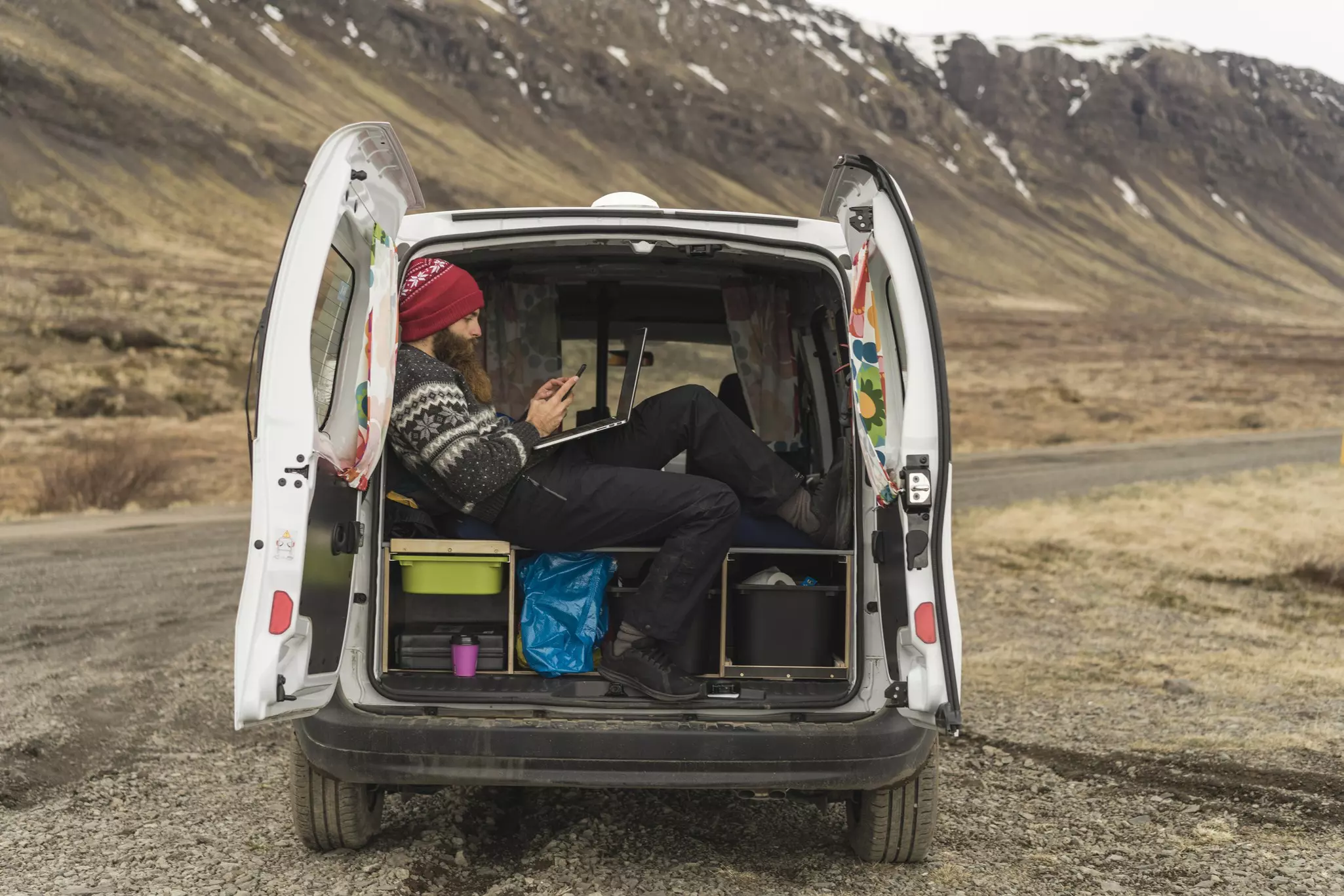 Iceland, man sitting in van using smartphone and laptop