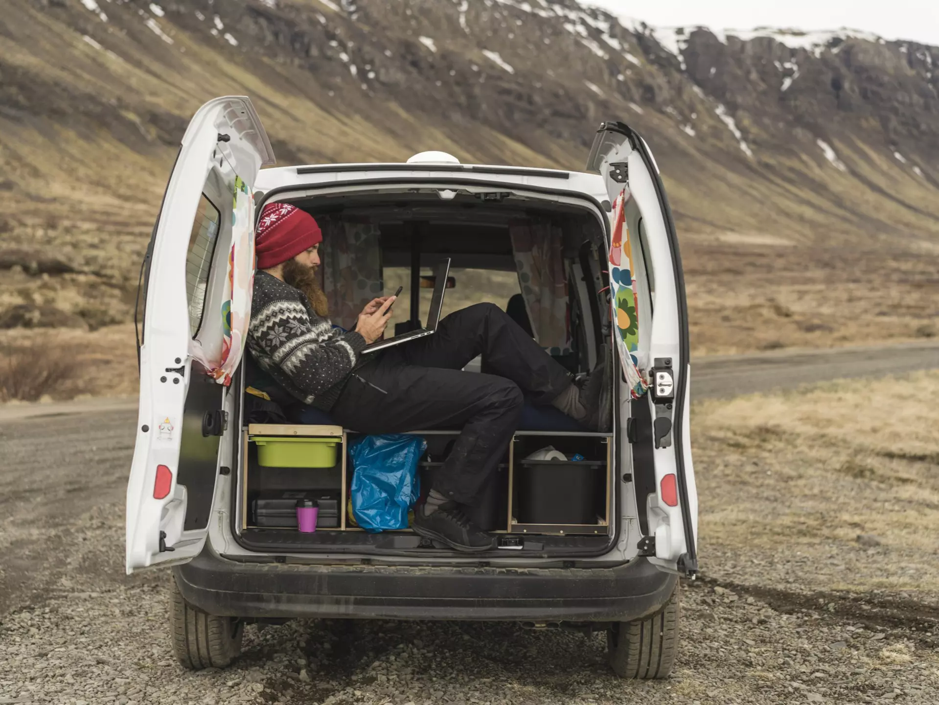 Iceland, man sitting in van using smartphone and laptop