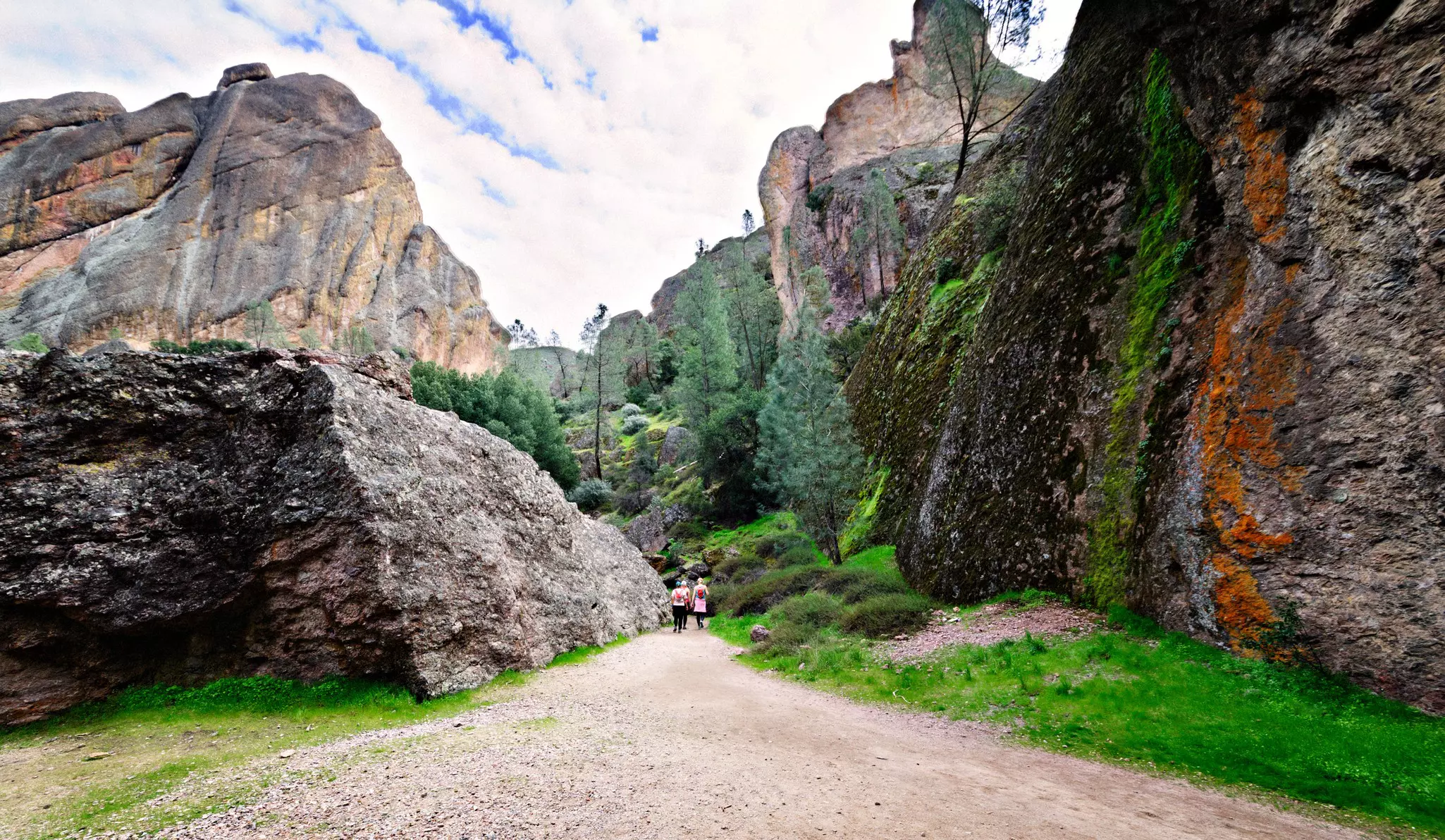 A small group of people follow a dusty trail between huge rocky boulders surrounded by trees