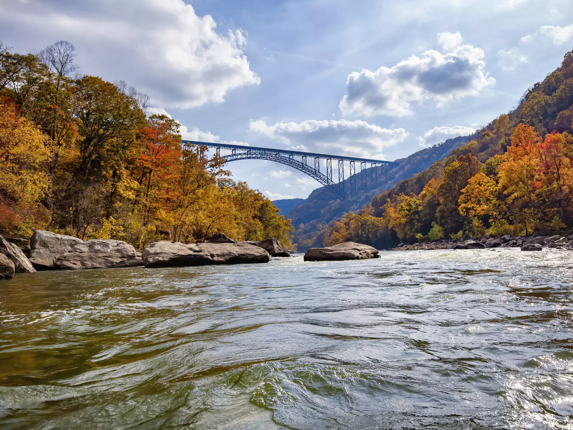 A river runs through a valley surrounded by trees changing into autumn colors of red, orange and gold. A large railway bridge passes above.