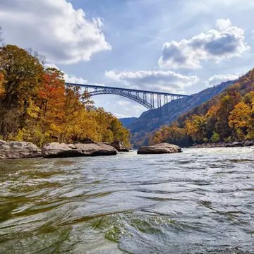 A river runs through a valley surrounded by trees changing into autumn colors of red, orange and gold. A large railway bridge passes above.