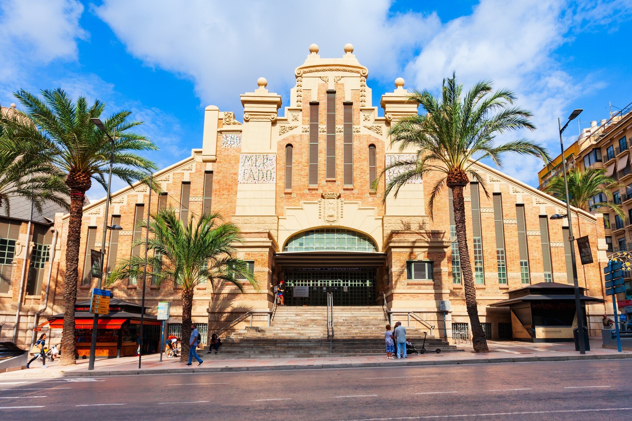 Ornate brick and cream stone market hall with a tiled 'Mercado' panel, wide entrance steps and palm trees under a blue sky