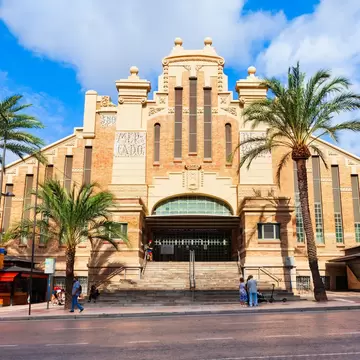 Ornate brick and cream stone market hall with a tiled 'Mercado' panel, wide entrance steps and palm trees under a blue sky