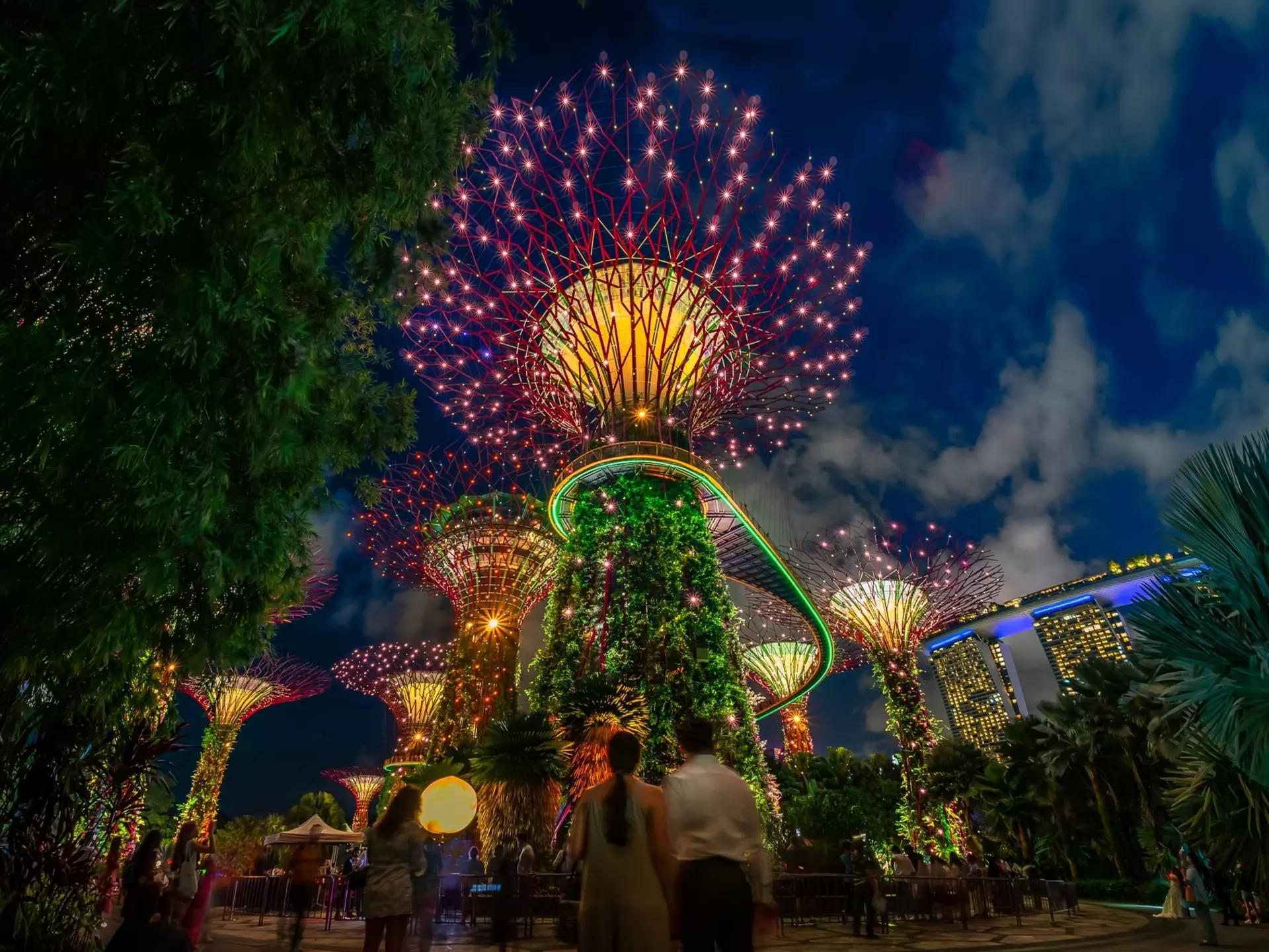People look up at a series of glass towers illuminated with bright lights after dark.