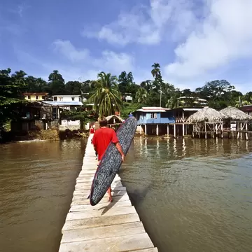 Man in a red shirt carrying a surfboard along a narrow wooden dock toward waterfront stilt houses and palm trees.