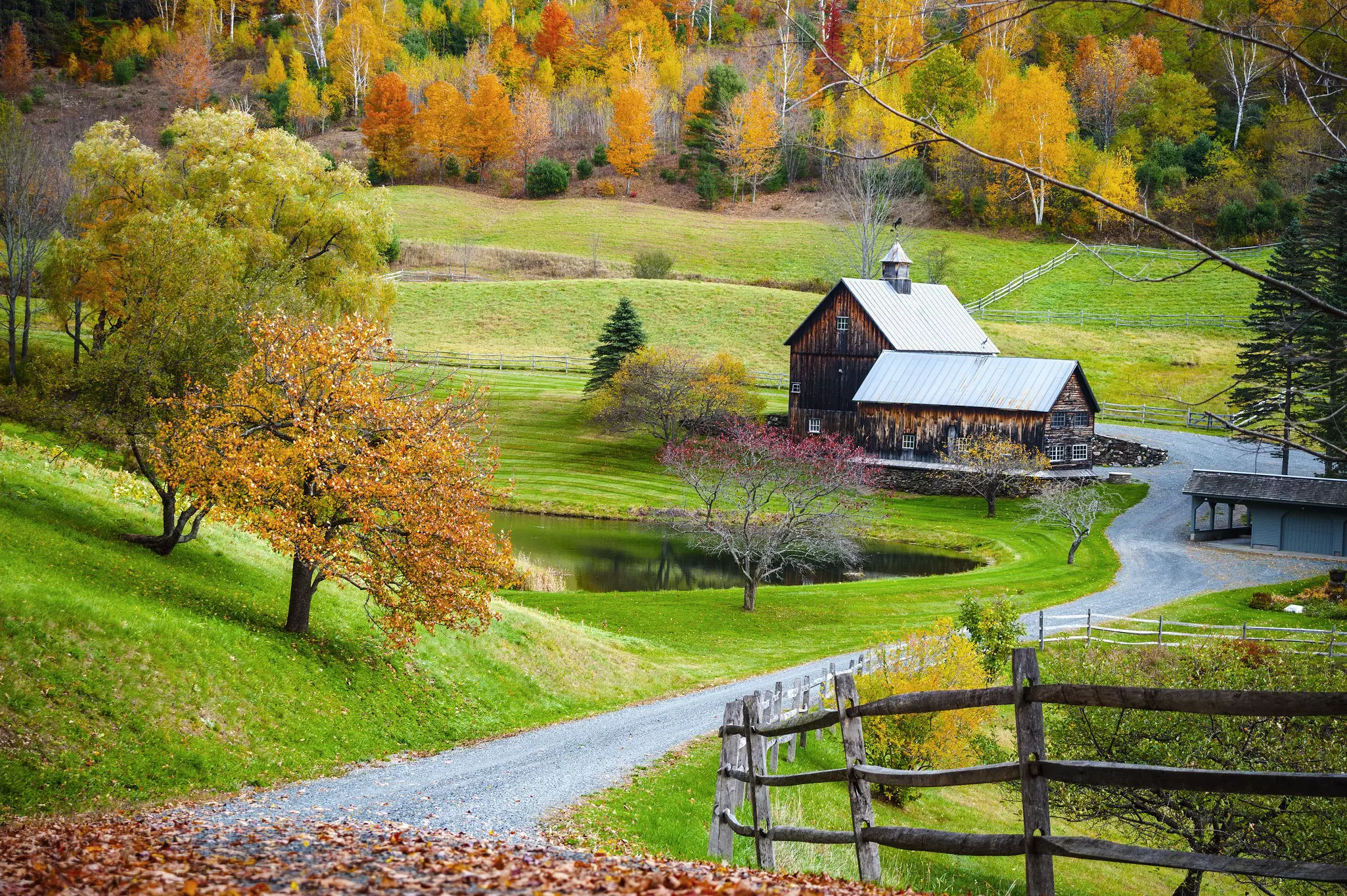 An unpaved road leads to a historic wooden barn surrounded by green lawns and trees bearing fall foliage