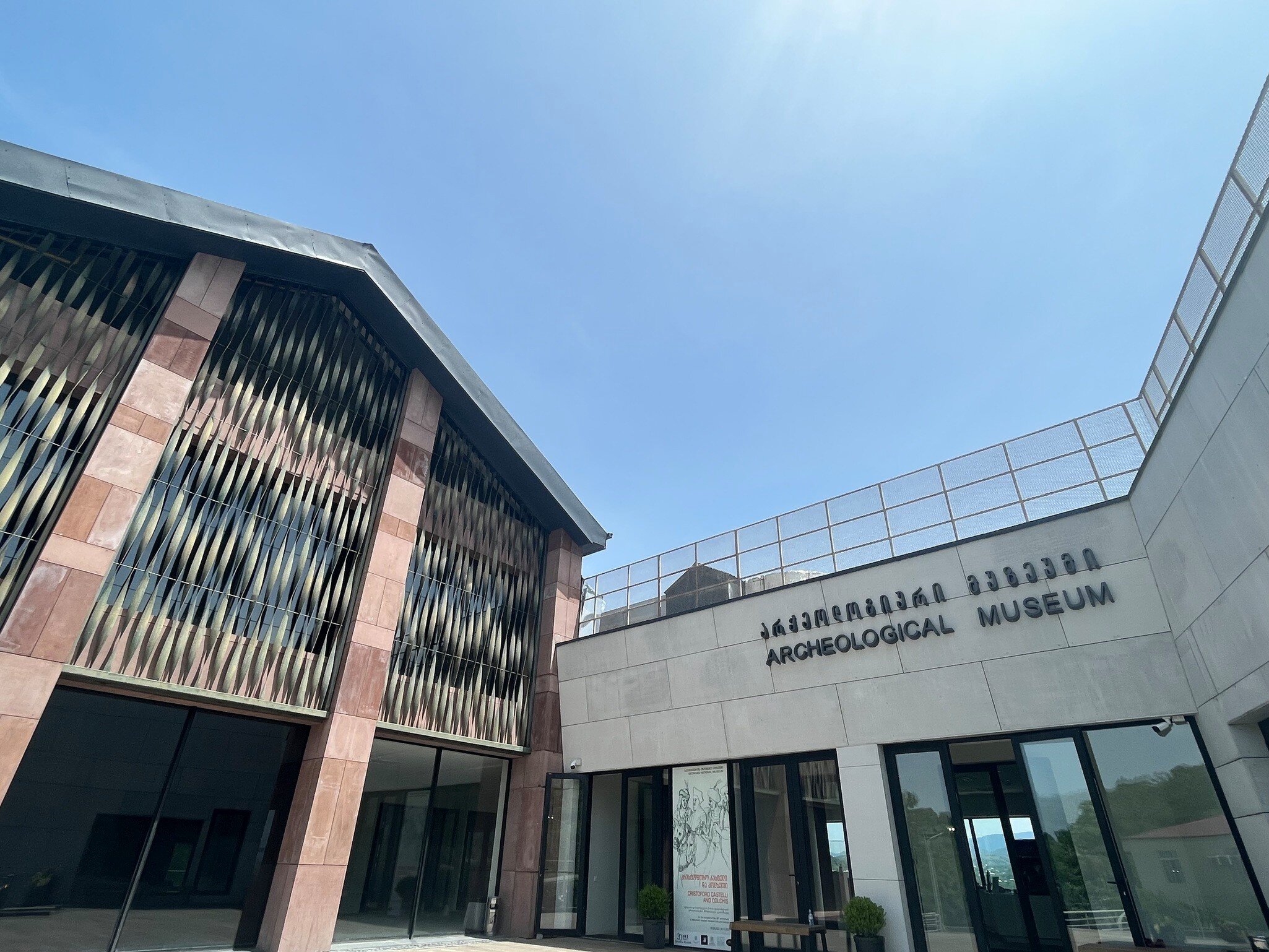 Modern museum with a red stone and metal lattice facade beside a pale wing signed Archeological Museum