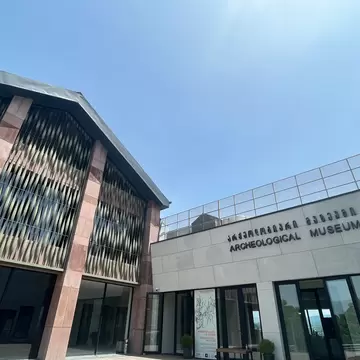 Modern museum with a red stone and metal lattice facade beside a pale wing signed Archeological Museum