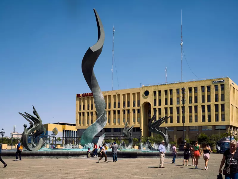 A sculptural fountain sits in a big square in front of a rectangle yellow building 