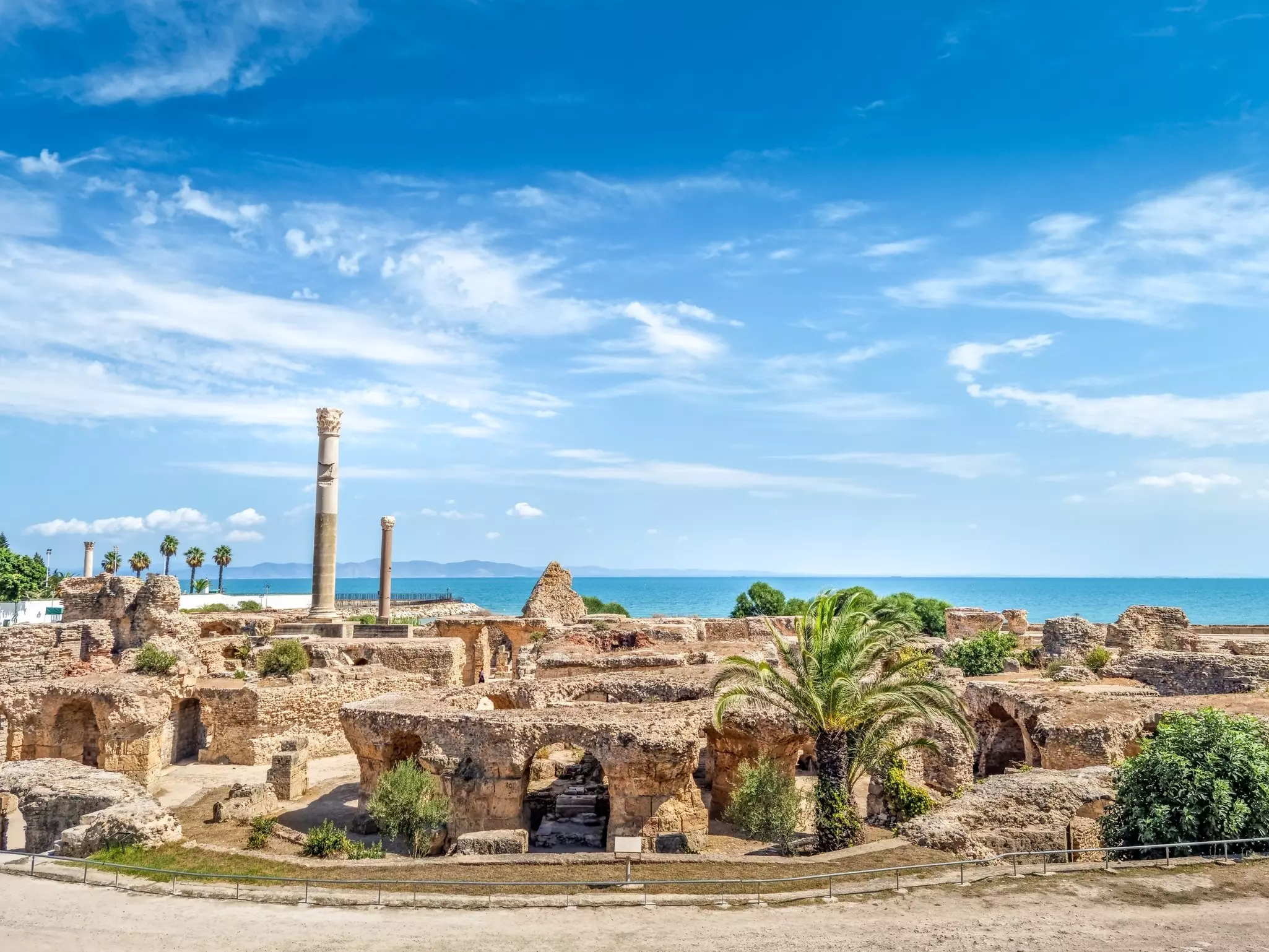 A series of ruined walls, doorways and tall columns among palm trees in a coastal location.