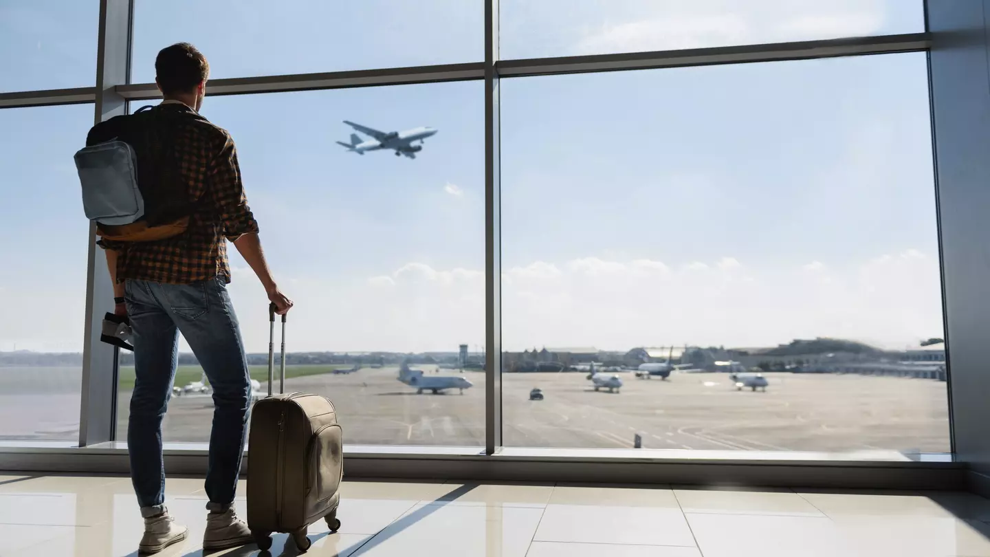 Young man is standing near window at the airport and watching plane before departure.