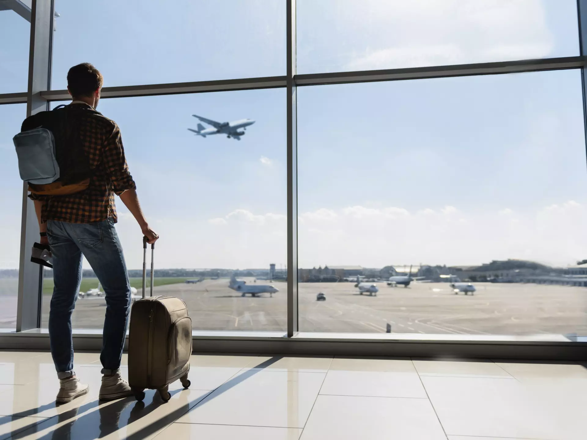 Young man is standing near window at the airport and watching plane before departure.