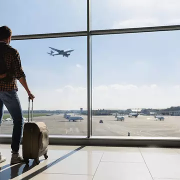 Young man is standing near window at the airport and watching plane before departure.
