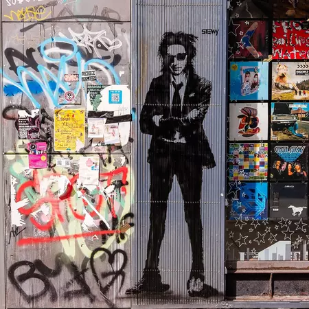 Close-up of building wall with graffitti lettering and a full-body stencil spray-painted in black of a man in a suit with pulled-down tie, ruffled long hair, and sunglasses.
