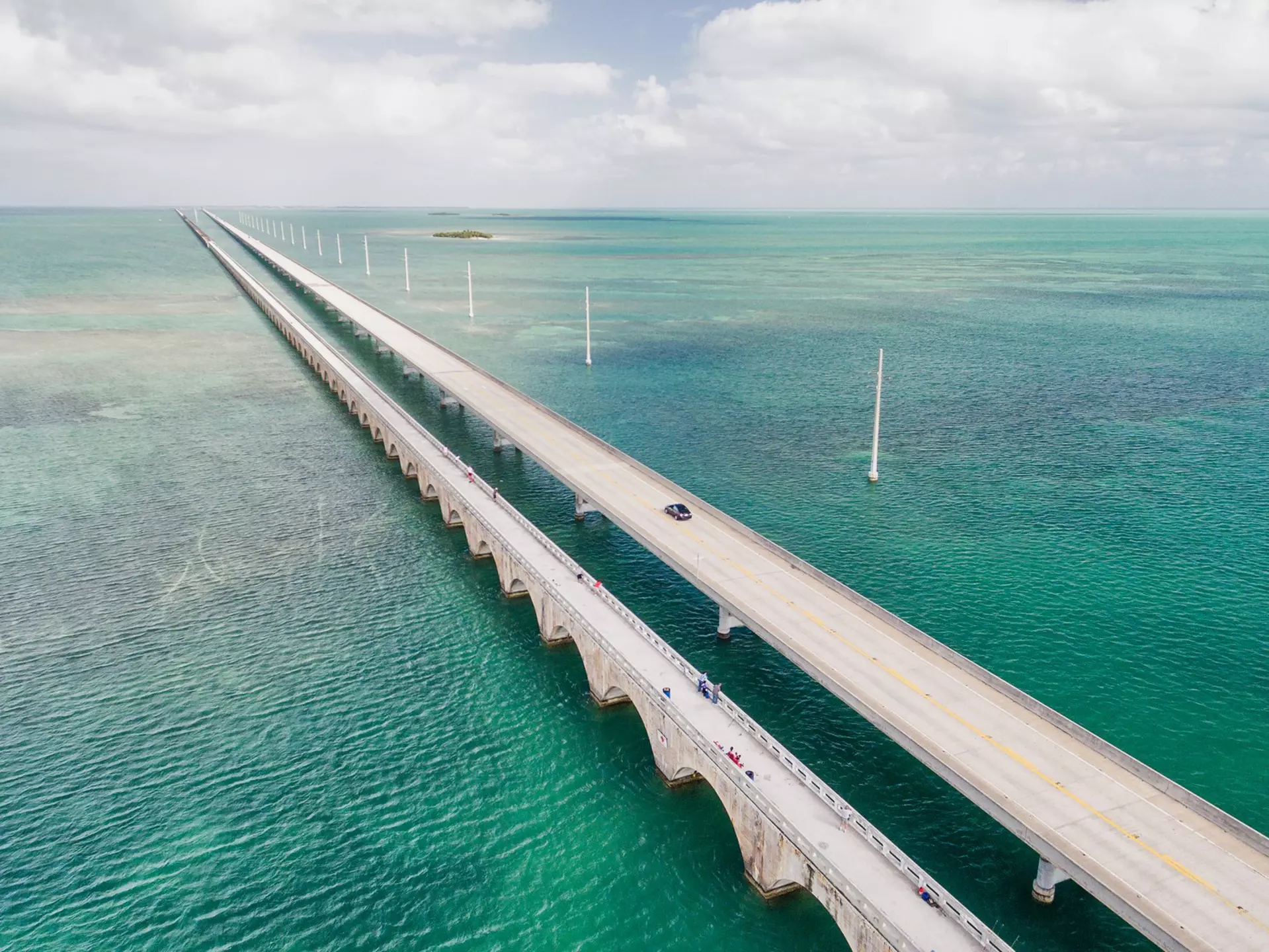 An overhead view of a long causeway crossing shallow water in many hues of blue and green.