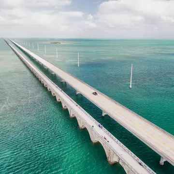 An overhead view of a long causeway crossing shallow water in many hues of blue and green.
