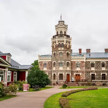 A granite manor with a tall crenellated tower, flanked by a red wooden building and a curving path across lawns under cloud
