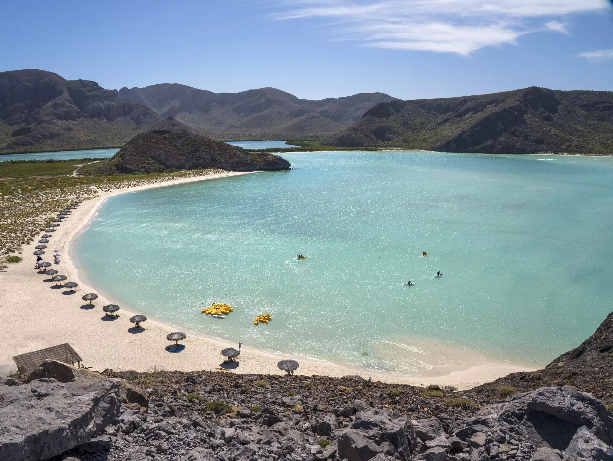 Aerial view of curved bay with light blue water on a sunny day. The sandy beach is lined with umbrellas in a row, and mountains are seen in the distance.