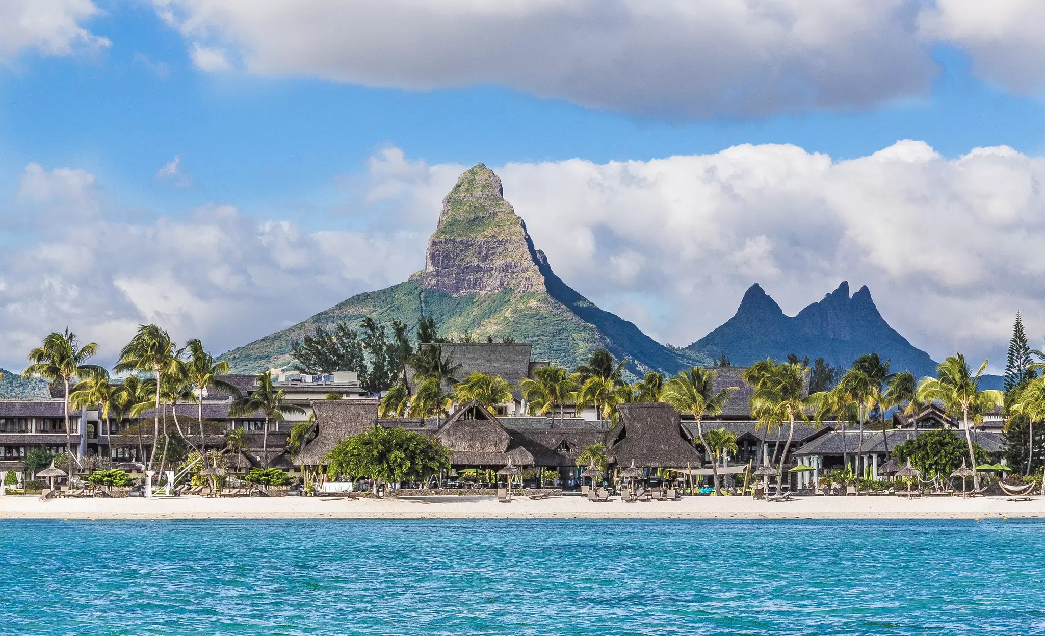 A gorgeous sandy beach lined with palm trees and a resort, backed by a huge pointed mountain 