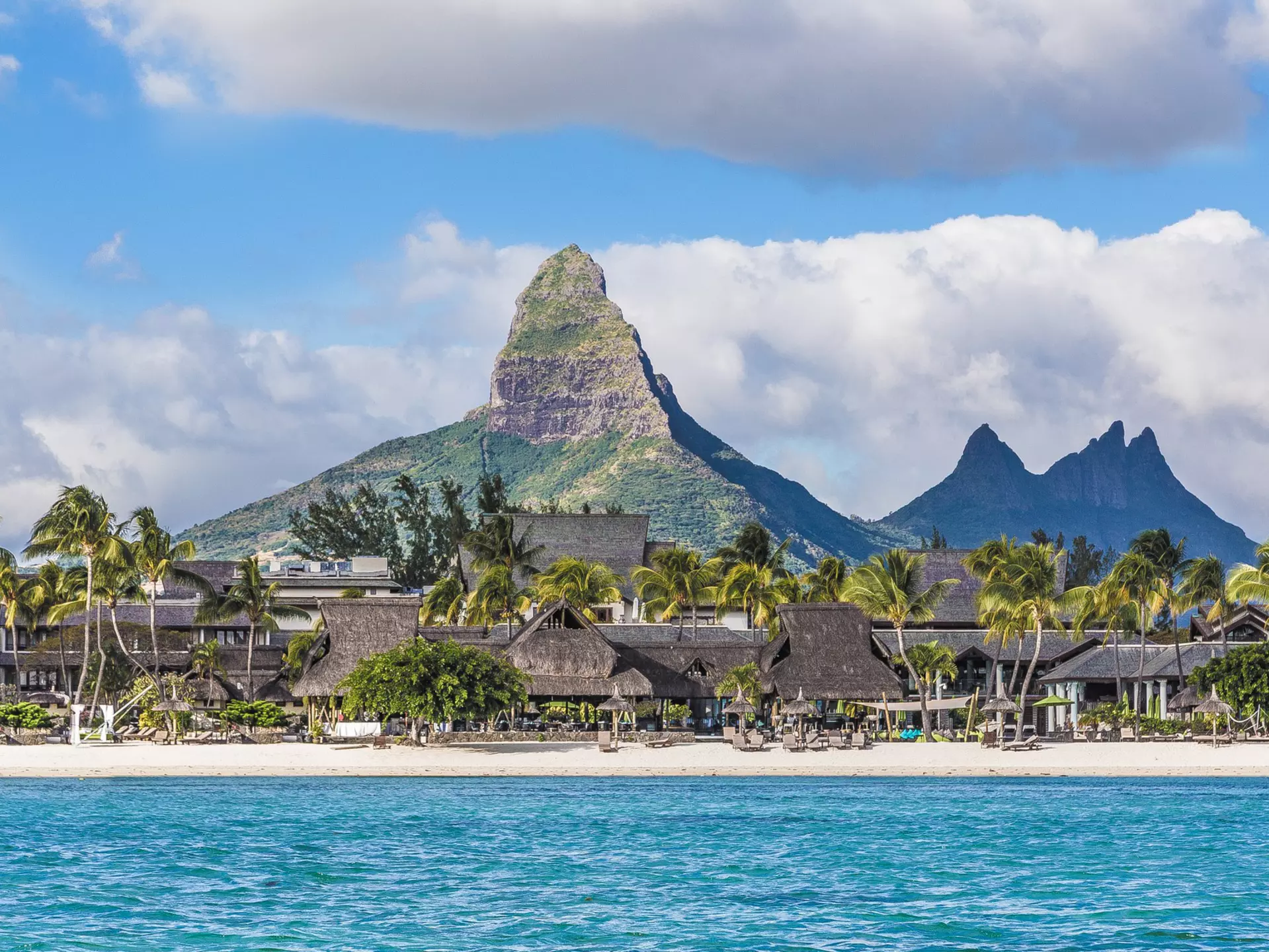 A gorgeous sandy beach lined with palm trees and a resort, backed by a huge pointed mountain 